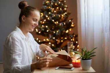 Beyaz gömlekli gülümseyen bir kadının yan görünüm portresi arka planda Xmas ağacıyla birlikte oturuyor, arkadaşı veya kocası için hediye kutusu hazırlıyor, yeni yıl hediyesini vereceği anı hayal ediyor..