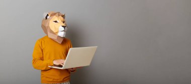 Aslan maskeli meşgul bir adamın portresi günlük tarz turuncu kazak giyiyor. Gri arkaplanda poz veriyor ve reklam için fotokopi alanı yaratıyor. Dizüstü bilgisayarda çalışıyor..