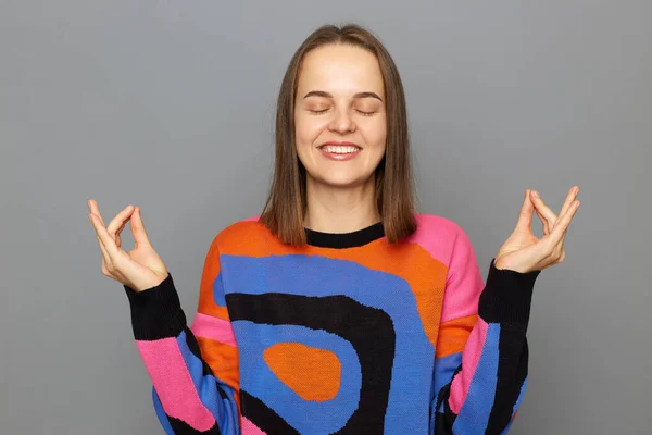Portrait of smiling satisfied woman with brown hair wearing colorful jumper posing isolated over gray background, standing and practicing yoga, smiling with satisfied expression.