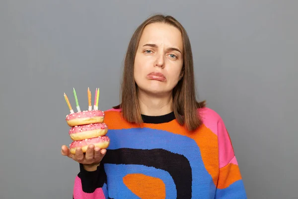 Portrait of sad unhappy young adult woman with wearing warm sweater holding donut with candles isolated over gray background, celebrating her birthday alone, looks with pout lips.