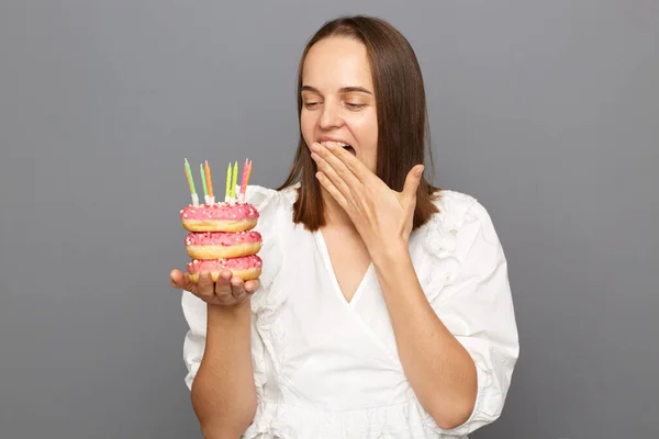 Portrait of surprised woman with brown hair wearing white blouse holding donut with candles isolated over gray background, covering mouth with palm, pleasant surprise.