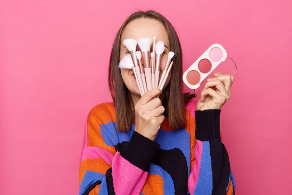 stock image Photo of anonymous funny woman with dark straight hair wearing jumper standing isolated over pink background, covering her face with cosmetics brushes, holding eyeshadow palette.