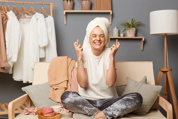 Image of hopeful smiling positive woman in towel on her head with rack clothes in living room, making wish, crossing fingers, hopes she finds what to wear.