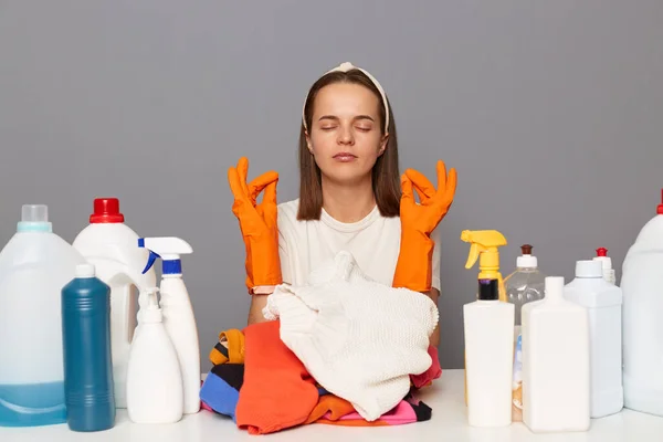 Indoor shot of woman wearing protective gloves, surrounded with many cleaning products, feels fatigue while cleaning house, trying to calm down and rest doing yoga and meditate.