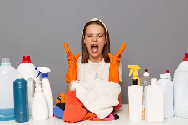 Image of hesitant angry woman with despair expression, sitting among different detergent, sitting at table with chemical products for tidying up, screaming with angry isolated over gray background.
