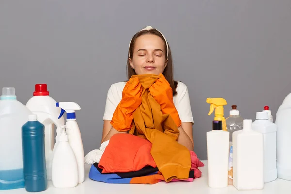 Image of attractive woman wearing headband, protective rubber gloves, surrounded with cleaning accessories, poses at desk, smelling fresh clothing after laundry, isolated over gray background.
