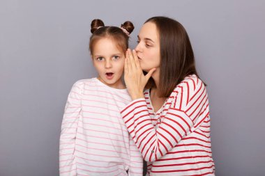 Annesinin, ağzı açık duran kızıyla, gri arka planda soyutlanmış günlük giysiler giyen kızıyla fısıldaşırken çekilmiş kapalı çekimler..
