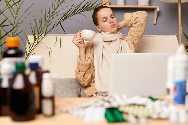 stock image Indoor shot of tired exhausted bored woman sitting on sofa wrapped in scarf surrounded with medicines, drinking tea, holding cup, touching her neck, looking at laptop display, posing in living.