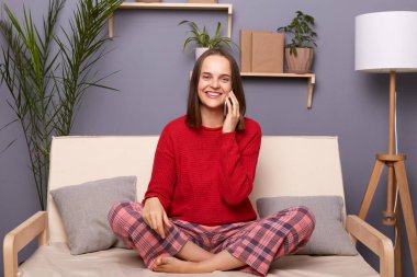 Kırmızı kazak ve kareli pantolon giyen, akıllı telefonla konuşan, hoş sohbetler yapan, evdeki koltukta oturan sevimli, genç bir kadının görüntüsü..