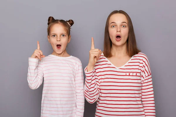 Horizontal shot of amazed excited mother and little daughter wearing casual style shirts, raised their fingers up, having idea, keeps mouth widely open, posing isolated over gray background.