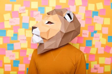 Turuncu kazak ve kağıt aslan maskeli, kendinden emin güçlü bir adamın kapalı çekimleri, güçlü görünüyor, başka yöne bakıyor, sarı arka planda izole edilmiş poz veriyor..