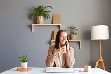 Güzel, gülümseyen, tatmin olmuş, resmi tarz ceket giyen, ofiste oturan, cep telefonuyla konuşan ve dinlenirken kahve içen bir kadın..