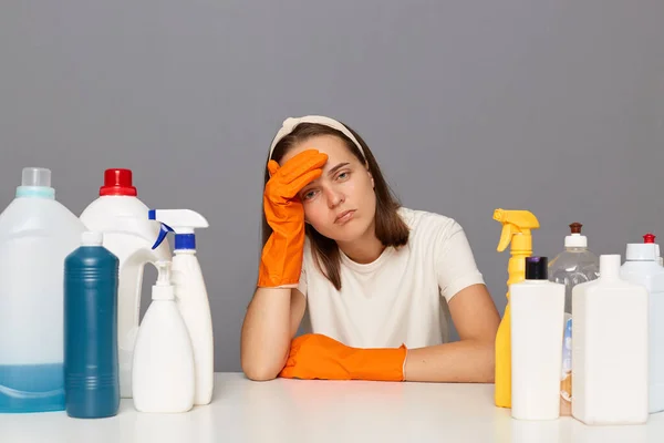 Unhappy woman with displeased expression, being tired, has fatigue look, wearing white t shirt sitting isolated over gray background, having headache, surrounded with cleaning chemical supplies.
