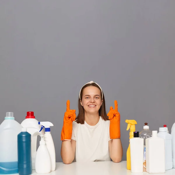 Woman wearing white t shirt and rubber gloves, pointing up at advertisement area, copy space for promotion, sitting isolated over gray background, being surrounded with cleaning detergents.