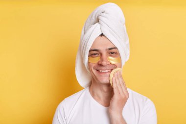 Göz altı yamalı, beyaz tişörtlü, sarı arka planda izole bir şekilde havluya sarılmış, elinde sünger tutan çekici ve pozitif bir adamın yatay çekimi..