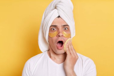 Şok olmuş beyaz tişört giyen, havluya sarılmış, yüzündeki kremi süngerle sıyırmış, sarı arka planda tek başına duran adamın görüntüsü..