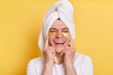 Son derece mutlu bir adamın portresi beyaz tişört giyiyor ve sarı arka planda izole bir şekilde havluya sarılı duruyor, süngeri elinde tutuyor, cildine bakarken gülüyor..