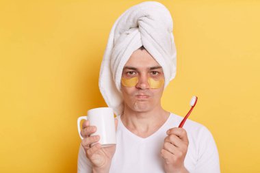 Gözlerinin altında yamaları olan, beyaz tişört giyen ve havluya sarılmış, sarı arka planda tek başına duran, elinde diş fırçası ve fincanı tutan kızgın üzgün adamın portresi..