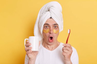 Şaşırmış bir adamın portresi, beyaz tişört giyen, sarı arka planda tek başına duran, diş fırçalayan, hijyen prosedürleri yapan, havluya sarılı şaşırmış bir adamın portresi..