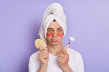 Gözlerinin altında yamalar olan, beyaz tişört giyen, havluya sarılı, mor arka planda tek başına duran, elinde sünger ve masaj rulosu tutan üzgün bir adamın portresi..