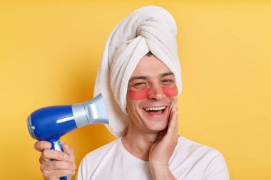 Beyaz tişört giyen, havluya sarılmış, sarı arka planda tek başına duran, elinde saç kurutma makinesi tutan iyimser adamın kapalı çekimleri..