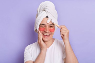Beyaz tişört giyen, havluya sarılı, mor arka planda yalnız duran alnına masaj yapan, cilt bakımı prosedürleri yapan bir adamın portresi..