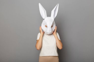 Horizontal shot of woman wearing white t shirt and paper rabbit mask standing isolated over gray background, taking off her mask or covering ears, mind blowing. clipart