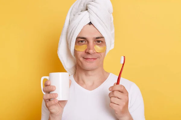 Hygiene, morning routine and teeth whitening. Calm handsome man brushes teeth, wears towel on head, holding white cup in hands. Stomatology, everyday life.