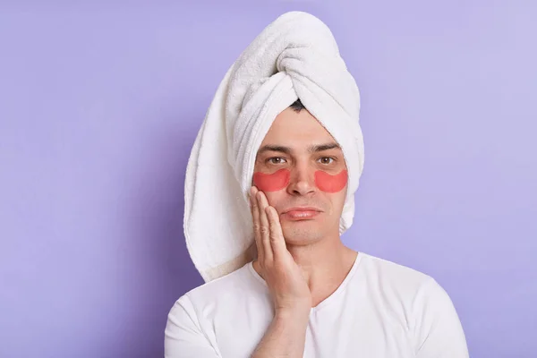 Horizontal shot of sick unhappy man wearing white T-shirt and being wrapped in towel standing isolated over purple background, posing with patches under eyes, touches cheek.