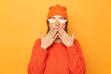 Şok olmuş turuncu kazaklı, bereli ve gözlüklü kadının sarı arka planda, ağzı palmiyelerle kaplanmış vaziyette dikilirken çekilmiş yatay görüntüsü..