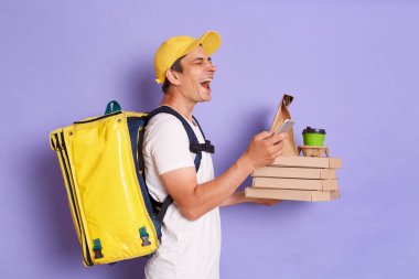 Heyecanlı çılgın kuryenin profil portresi elinde pizza kutuları ve mor arka planda izole edilmiş kahve tutuyor, cep telefonu kullanıyor, müşteriyle iletişim kuruyor..