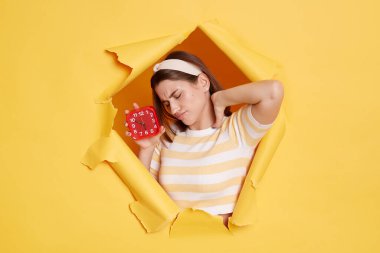 Sağlıksız, yorgun, çizgili tişört ve saç bandı takan, sarı duvardaki kağıt delikten geçen, alarm saatini gösteren, boynundaki acıyı hisseden bir kadının fotoğrafı..