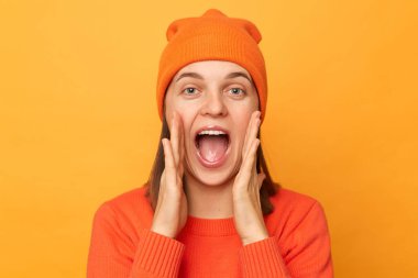 Kapalı alanda, turuncu süveter ve şapka giyen, sarı arka planda izole edilmiş, çığlık atan ve ilan veren genç, hippi bir kadının fotoğrafı..