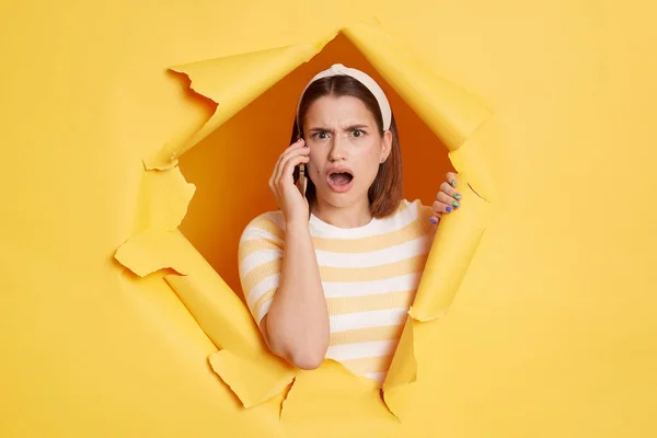 Portrait of surprised young woman wearing striped T-shirt and hair band, looking at camera with big eyes and talking on cell phone, breaks through yellow paper background in ripped hole.