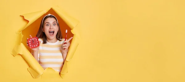 Amazed smiling woman wearing striped T-shirt and hair band, breaking through paper hole in yellow wall, holding alarm clock and toothbrush in hands, time to clean teeth, copy space for advertisement.