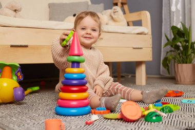 Evde piramit ve oyuncaklarla oynayan, sıcak bej kazak giyen, oturma odasındaki kanepenin yanında oturan mutlu, pozitif, güzel bir bebek resmi..