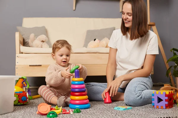 Indoor shot of kid playing with pyramid toy together with mother, happy little girl collects a toy pyramid of rings on floor in a kindergarten, development of fine motor skills in children lifestyle.