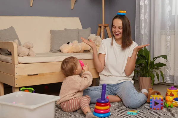 Image of positive laughing mother wearing white shirt and jeans playing with her little daughter at home while sitting on floor, holding rings of pyramid on her head.