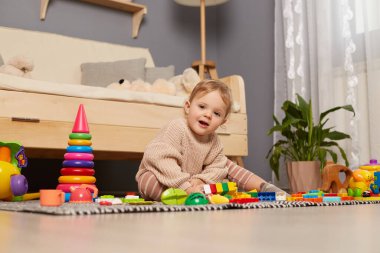 Bej kazak giyen, renkli oyuncaklarla çevrili, kameraya bakan, evde oynayan, mutlu bir şekilde gülümseyen sevimli bir bayan çocuk portresi..