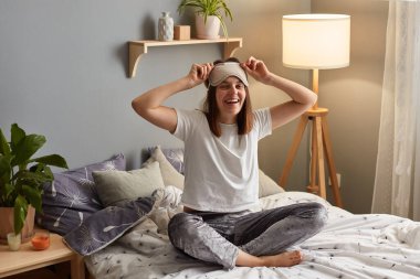 Yatakta rahat bir yatak odasında rahat bir şekilde oturan, neşeli bir yüz ifadesiyle kameraya bakan, son derece mutlu bir kadın fotoğrafı..