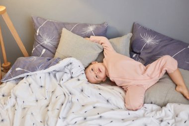 Yatak odasında, tuhaf bir ifadeyle uzanmış, yatakta tembellik edip yastıklarda poz veren sevimli, sevimli bir kız bebek resmi. Mutlu çocukluk.