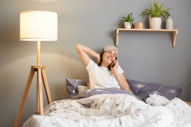 Memnuniyet dolu, gözbağı takan, iç kısımda yatağında oturan, rahat bakışlı, akıllı bir telefonla konuşan, dostça gülümseyen kadın portresi..