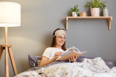 Evde, beyaz t-shirt giyen, evde kitap okuyan, ilginç romanlardan hoşlanan, mutlu bir ifadeyle sayfalara bakan, mutlu bir yetişkin kadın resmi..