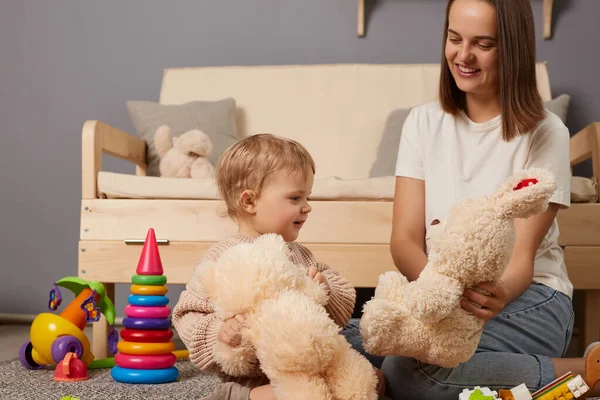 Indoor shot of smiling positive woman and her daughter playing together having fun together, playing soft toys at home on the floor, family play with soft teddy bear and rabbit.