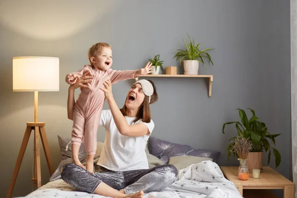 Image of joyful cheerful mother with her baby sitting o bed and having fun together, mommy and daughter playing in bedroom, kids spreads arms like flying.