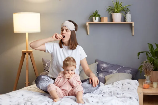Indoor shot of sleepy exhausted woman wearing pajama and blindfold with her baby daughter in the bedroom sitting on bed, playing with toddler kid, yawning, covering mouth with palm.