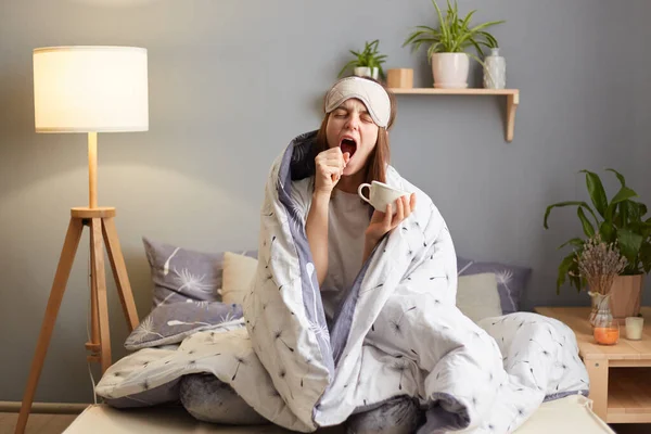 Indoor shot of sleepy woman wrapped in a blanket after waking up, holding cup of coffee, covering mouth with hand, feels sleepless, drinking energetic beverage.
