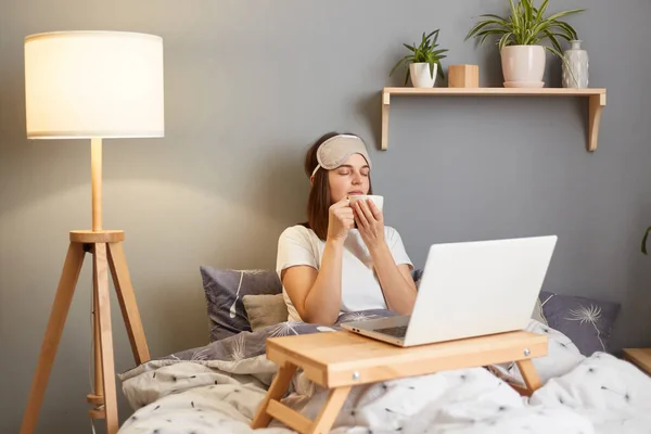 Indoor shot of satisfied delighted woman wearing sleep mask sitting with laptop in bed at home, smelling coffee with pleasant expression, working online on pc computer.
