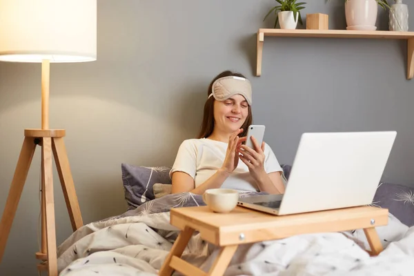 Photo of young adult brown haired woman wearing sleep mask sitting with laptop in bed at home and typing on mobile phone, writing message or scrolling online.