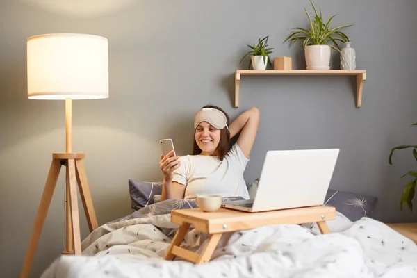 Horizontal shot of smiling attractive Caucasian woman in sleep mask sitting with laptop in bed at home and using smart phone, having positive facial expression, spending Sunday morning in bedroom.